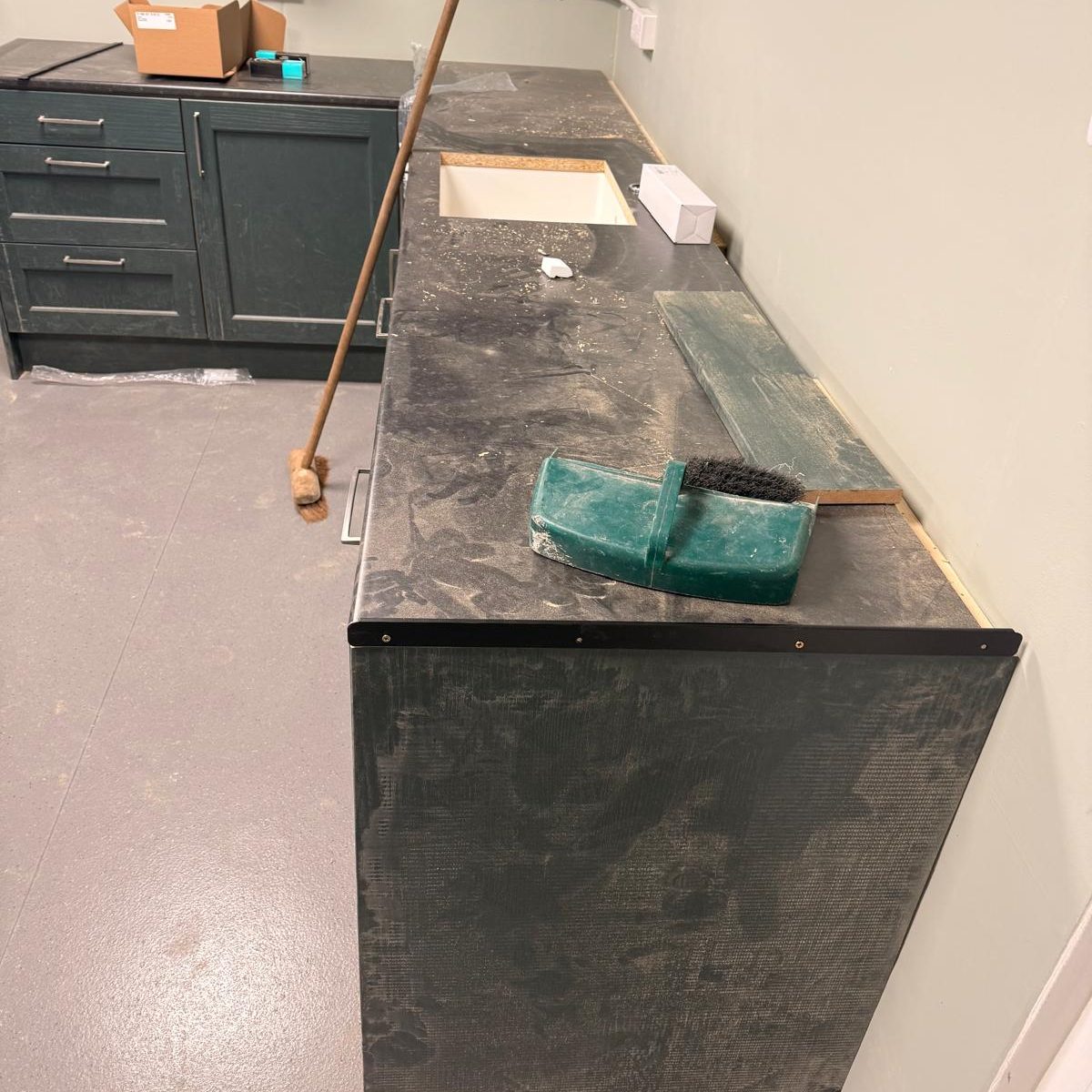 Commercial Deep Cleaning – Before Photo | Professional Results/ dirty sink & counter top A dusty dark countertop with a green cleaning tool and a sink area, against a grey wall.